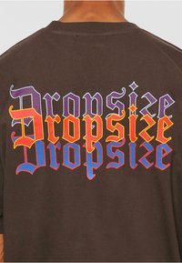 Brun bomulls-T-skjorte med flerfarget tekst som sier "Dropsize" i fet, stilisert skrift på baksiden.