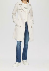Jasnobeżowy płaszcz typu trencz z dwurzędowym zapięciem, dużymi guzikami i bocznymi kieszeniami, noszony na paskowanej koszuli i niebieskich dżinsach z białymi trampkami.