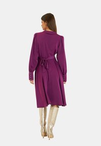 Lila Langarmkleid mit einem Gürtel, Knopfakzenten an den Ärmeln und ausgestelltem Rock; kombiniert mit glänzenden silbernen Stiefeln bis zum Knie.