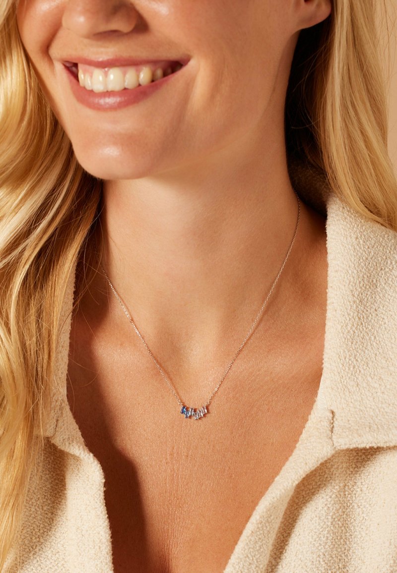 Zilveren ketting met kleine blauwe en heldere edelstenen in een gebogen lijn. Draagt op blote huid met een gestructureerd, lichtgekleurde shirt.