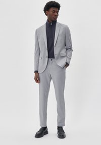 Ljust grå kostym med texturerad tyg, slim-fit design, enkelknäppt kavaj och matchande byxor. Tillsammans med en mörk zip-krage topp och skor.