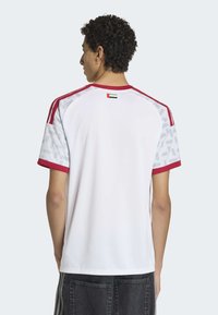 Chemise de sport blanche avec accents rouges et manches grises à motifs. Petit détail de drapeau à l'arrière du col. Manches courtes, col rond.