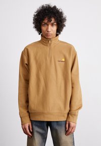 Brown fleece pullover with a high collar and quarter zip, featuring a logo in yellow and maroon on the left chest. Loose fit, ribbed cuffs.