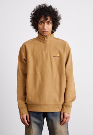 Sudadera de felpa marrón con cuello alto y media cremallera, que presenta un logo en amarillo y granate en el lado izquierdo del pecho. Corte holgado, con puños de canalé.