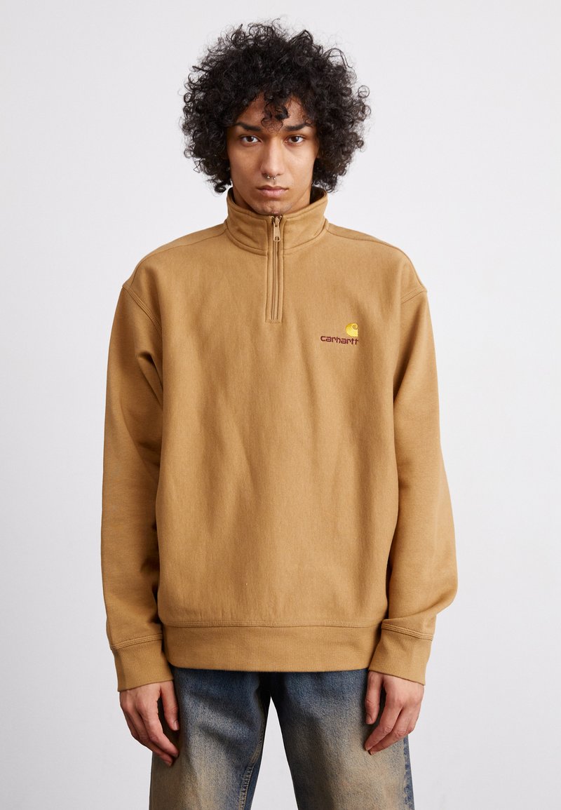 Brown fleece pullover with a high collar and quarter zip, featuring a logo in yellow and maroon on the left chest. Loose fit, ribbed cuffs.