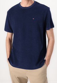 Mann trägt ein navyblaues kurzärmliges Hemd mit kleinem Logo auf der Brust, beiges Hemd, steht mit den Händen in den Taschen vor einem einfarbigen Hintergrund.