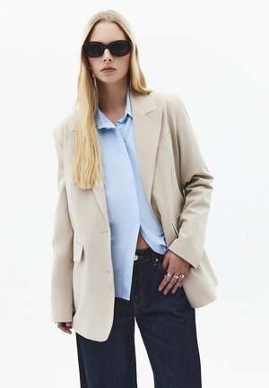 Lyshåret kvinde iført oversized beige blazer, lyseblå skjorte, mørke jeans, sorte solbriller og sølvringe mod en hvid baggrund.
