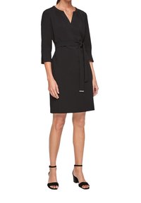 Svart klänning med V-ringning, trekvartslånga ärmar och knytmidja. Slät tyg, knälång nederkant, kombinerad med svarta remsandaler.