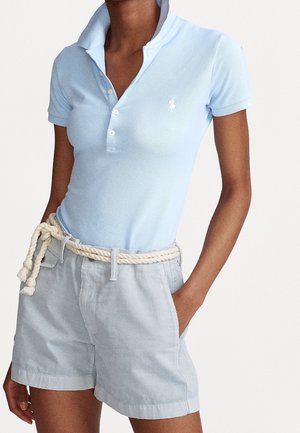 Vrouw draagt een lichtblauw poloshirt met korte mouwen met wit logo, lichtblauwe korte broek en een dikke geknoopte touwceintuur, hand in de zak.