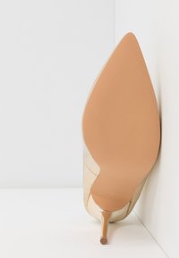 Chaussure à talon haut beige à bout pointu, texture lisse et talon fin, vue par en dessous sur un fond clair.