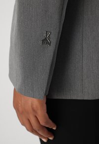 Gray blazer sleeve featuring a smooth texture, tailored fit, and a black metal emblem detail near the cuff.