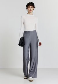 Una persona giovane è in piedi indossando una maglietta a maniche lunghe bianca a coste, pantaloni grigi a gamba larga, sneakers bianche e tiene in mano una giacca nera piegata.