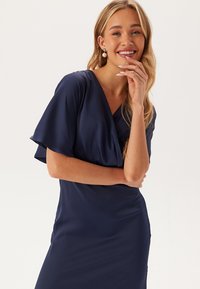 Námořnická modrá zavinovací šaty s krátkými volánovými rukávy. Vyrobené z hladké látky, mají hluboký výstřih do V a přiléhavý střih.