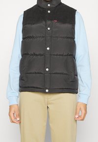 Chaleco negro acolchado con cuello de pana, botones a presión y bolsillos laterales. Llevado sobre una camisa de manga larga azul claro.