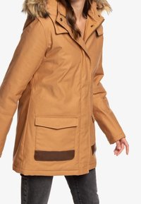 Chaqueta impermeable marrón con capucha forrada de pelo desmontable, que cuenta con bolsillos frontales y acentos de cuero marrón. Mangas largas y corte relajado.