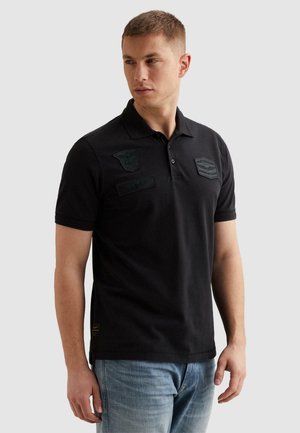 Man draagt een zwart poloshirt met geborduurde emblemen op de borst en lichtblauwe spijkerbroek, kijkt naar zijn linkerzijde.