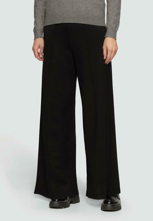 Person iført sorte bukser med vide ben, grå bluse med lange ærmer og sorte kraftige sko, stående med hænderne afslappet ned langs siden.