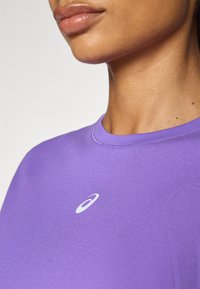Purple athletic shirt featuring a round neckline and a small white logo on the chest. Smooth texture and fitted design.