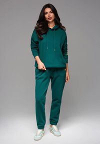 Sudadera de algodón verde con cordones y puños acanalados, combinada con joggers a juego. El modelo lleva zapatillas de color claro.