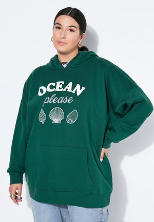 Frau mit dunklen Haaren, die einen grünen Hoodie mit dem Aufdruck „OCEAN please“ und drei weißen Muscheln trägt, posiert mit der Hand in der Hüfte vor weißem Hintergrund.