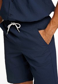 Marineblauwe mannen shorts in een gestructureerde stof, met een elastische tailleband en een witte trekkoord, evenals zijzakken.