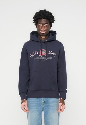 Marineblå hettegenser med frontlomme, snor i hetta og logo i rødt og hvitt. Kombinert med lyseblå jeans.
