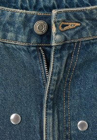 Pantalones vaqueros de mezclilla con cierre de cremallera estándar, botón de metal, costura color beige y detalles a presión en color plateado en la cinturilla.