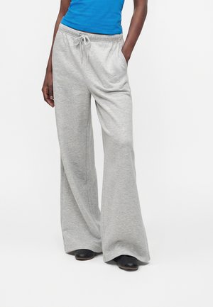 Person iført lysegrå vidde joggingbukser med snor i taljen og sorte sko, hånd i lommen, stående mod hvid baggrund.