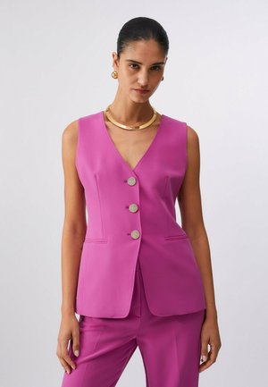 Donna che indossa un gilet senza maniche color magenta con bottoni e pantaloni abbinati, accessoriata con collana e orecchini d'oro, su sfondo uniforme.