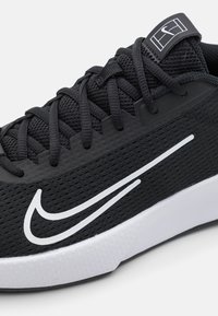 Zapatilla deportiva negra con parte superior de malla texturizada, detalle en color blanco del swoosh en contraste, cordones planos y suela de goma blanca de perfil bajo.