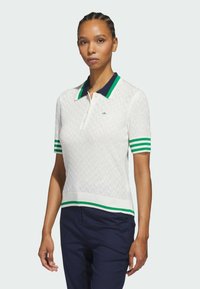 Hvit polo med et teksturert mønster, marineblå krage og grønnstripete ermer. Har en grønn logo på brystet og en tettsittende design.