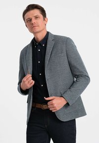 Grå vävd kavaj med skräddarsydd passform, slagkrage och påsydda fickor. Buren över en mörk knappad skjorta, i kombination med mörka jeans.