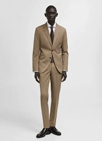 Abito beige con una leggera fantasia a righe, giacca monopetto e pantaloni abbinati. Accompagnato da una camicia bianca e una cravatta marrone. Scarpe eleganti marroni.