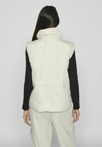 Fyllig, ärmlös väst i off-white med hög krage och sömda paneler, tillsammans med randiga byxor. Strukturerad svart långärmad tröja syns.