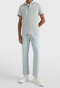 Mann, der ein hellgraues Poloshirt mit gestreiftem Kragen, hellblaue gemusterte Jeans und weiße High-Top-Sneaker trägt, steht mit einer Hand hinter dem Rücken.