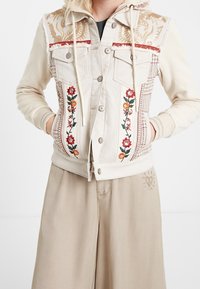 Chaqueta crema con bordados florales en los paneles frontales, mangas beige y capucha, combinada con pantalones beige que presentan un pequeño diseño de sol y corazón.