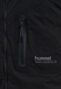 Schwarze technische Jacke mit einer mit Reißverschluss versehenen Brusttasche, glatter Stofftextur und einem in Grau auf der unteren rechten Seite gedruckten Logo.