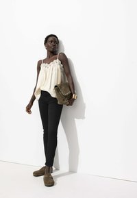 Camisole crème avec dentelle, associée à un jean skinny noir et des sabots en suède brun. Le mannequin tient un sac à bandoulière vert. Fond uni.