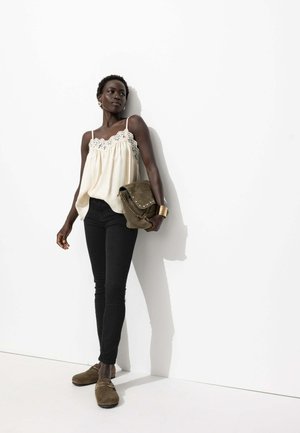Camisole crème avec dentelle, associée à un jean skinny noir et des sabots en suède brun. Le mannequin tient un sac à bandoulière vert. Fond uni.