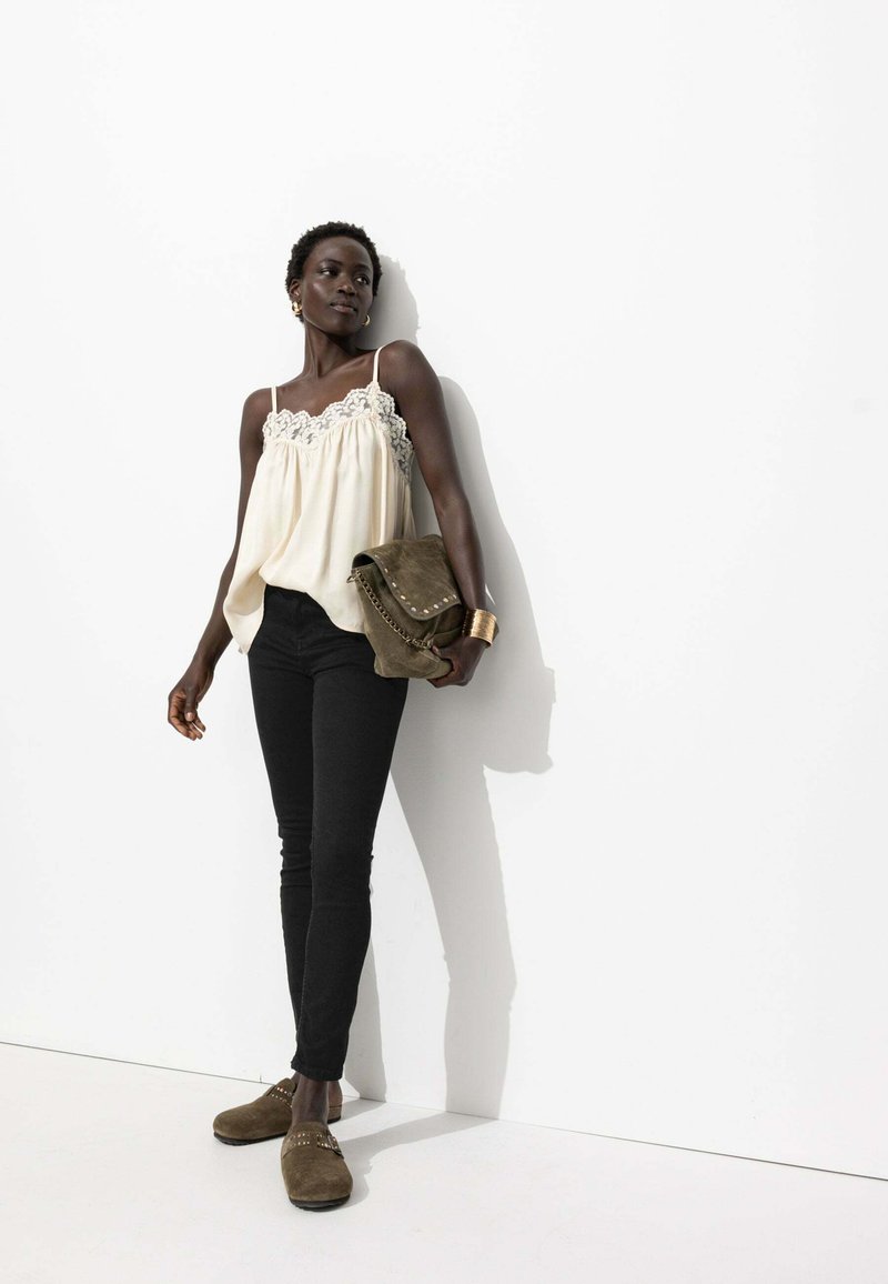 Camisole crème avec dentelle, associée à un jean skinny noir et des sabots en suède brun. Le mannequin tient un sac à bandoulière vert. Fond uni.