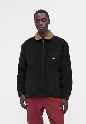 Homme portant une veste noire zippée avec un col beige et une poche avant, associée à un pantalon rouge, debout devant un fond blanc uni.