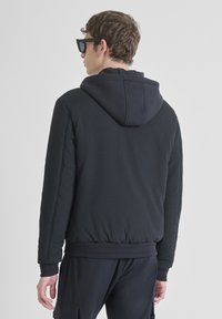 Man in een zwart gestructureerd jack met capuchon en zwarte broek, gezien vanaf de achterkant, tegen een effen lichte achtergrond.