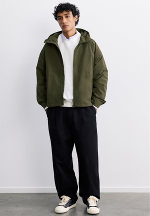 Ung mand iført grøn hættetrøje, hvid sweatshirt, sorte bukser og sort-hvide sneakers står op ad en ensfarvet hvid væg.