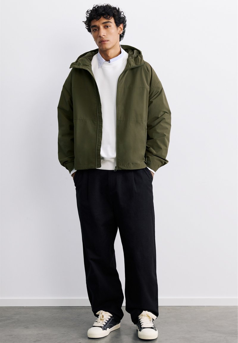 Jeune homme portant une veste à capuche verte, un sweat-shirt blanc, un pantalon noir et des baskets noires et blanches, debout contre un mur blanc uni.