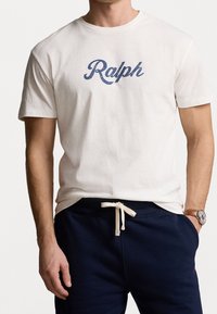 White cotton T-shirt featuring "Ralph" in navy blue script. Short sleeves, crew neck design, and relaxed fit. No notable embellishments.