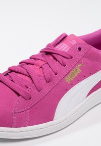 Baskets en daim rose avec accents blancs, équipées de lacets plats, d'un col rembourré et d'un logo doré sur le côté. Extérieur texturé et semelle lisse.