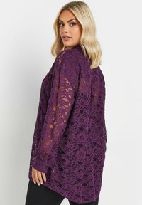 Purple lace shirt with long sleeves, floral pattern, and a relaxed fit. Features sheer texture and a curved hem.