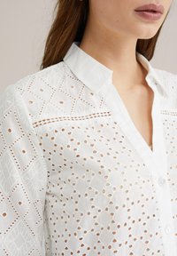 Camicia bianca con bottoni, realizzata in tessuto testurizzato con motivi a diamante e dettagli a taglio, dotata di colletto a punta e una patta dei bottoni visibile.