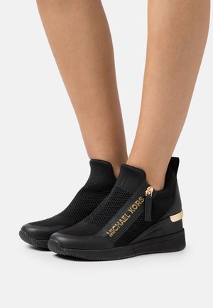 Baskets hautes noires sans lacets avec tiges en maille, logo doré "MICHAEL KORS", fermetures éclair latérales et accents dorés au talon, portées sur jambes nues.