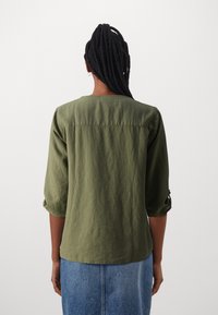 Olivengrünes, lockeres Shirt mit drei Viertelärmeln, strukturiertem Stoff und Rundhalsausschnitt. Zu den Details gehören umrollbare Ärmel und ein gerader Saum.